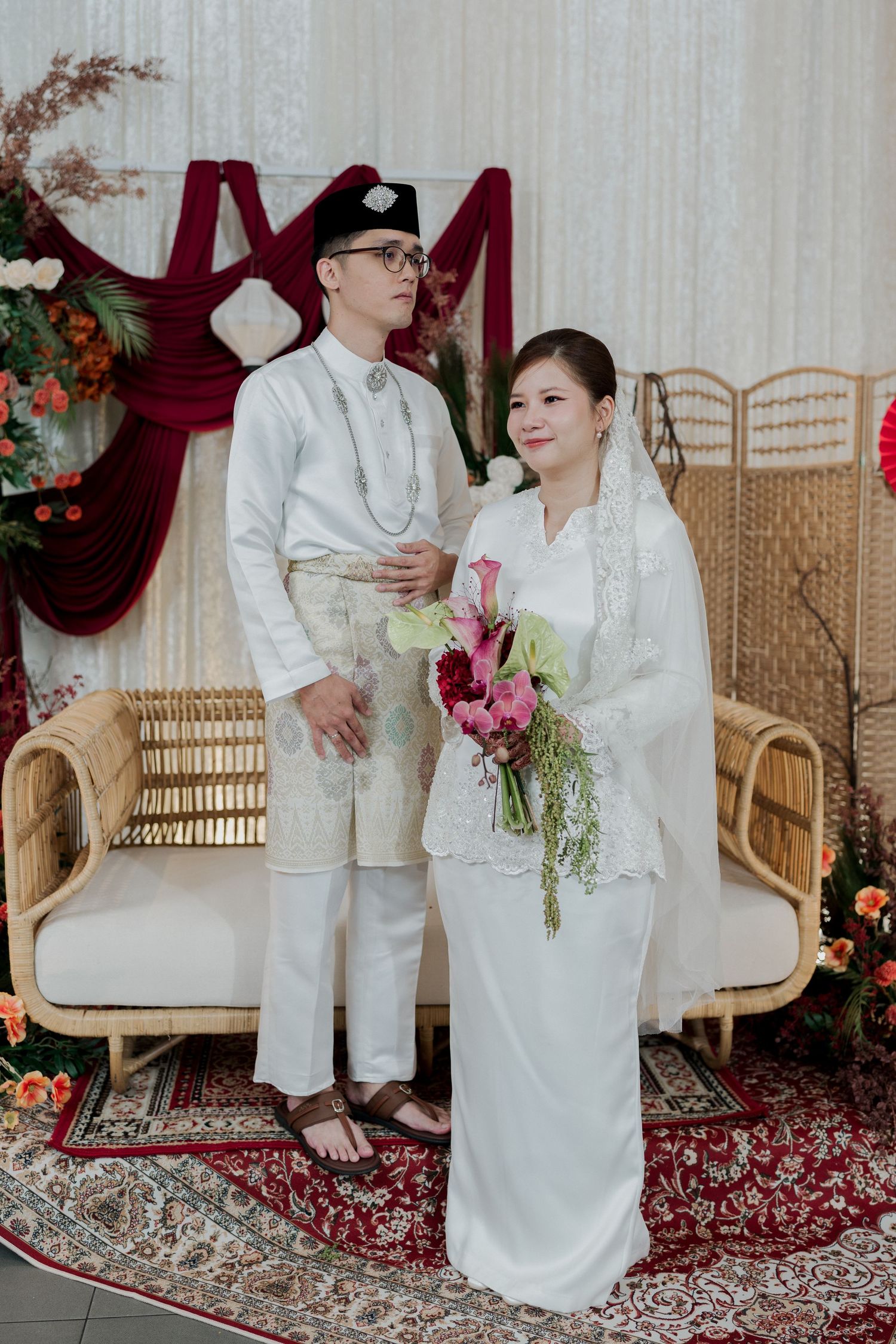 A Malay couple in traditional white wedding attire poses with a floral bouquet at an elegant decorated ceremony venue.