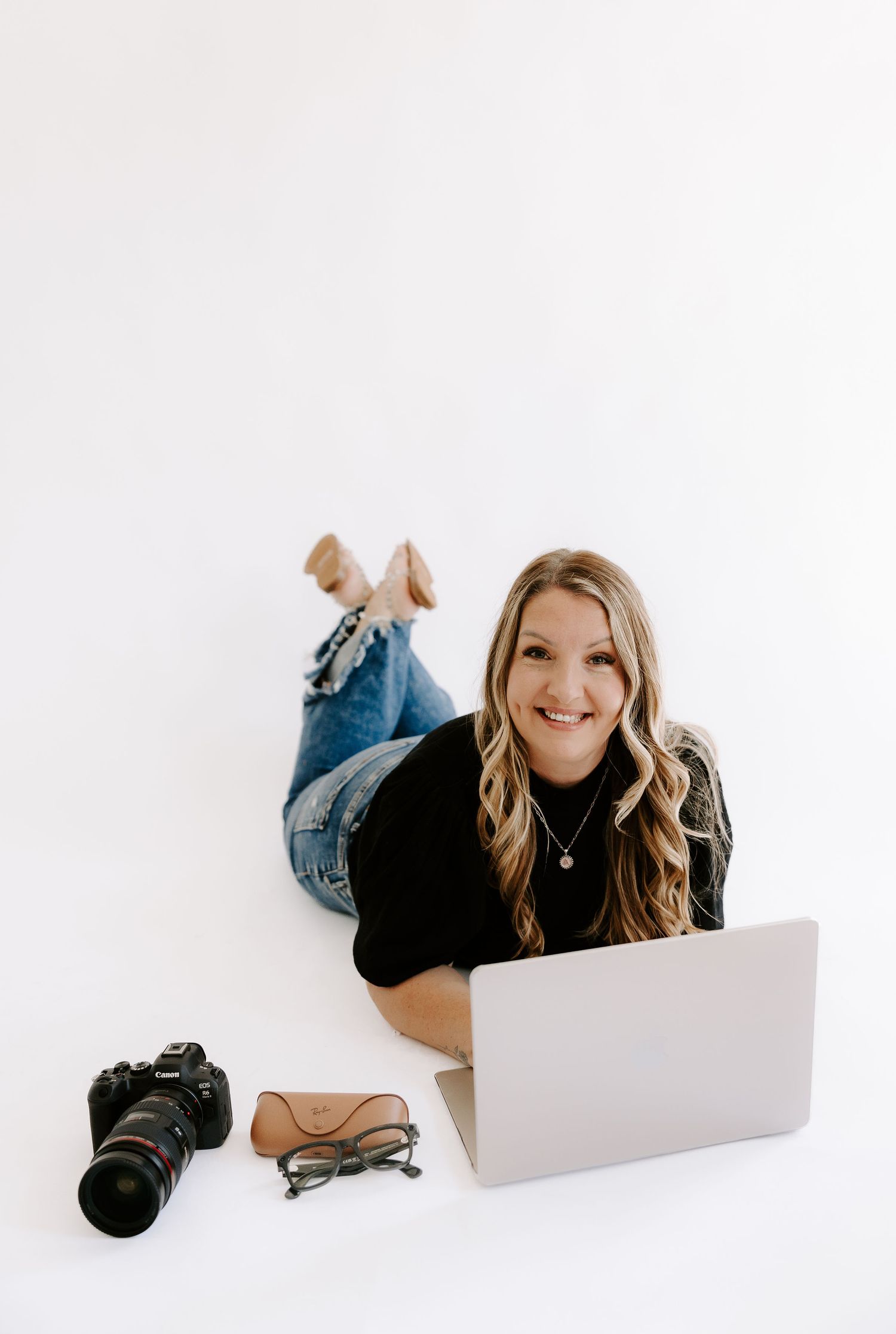Smiling blonde woman lying on white floor with laptop, DSLR camera, and glasses in a casual black top and jeans.