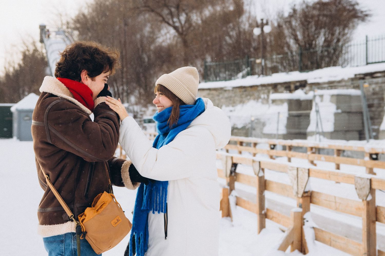 Two women in winter coats share a warm moment outdoors in a snowy setting, one adjusting the others scarf.