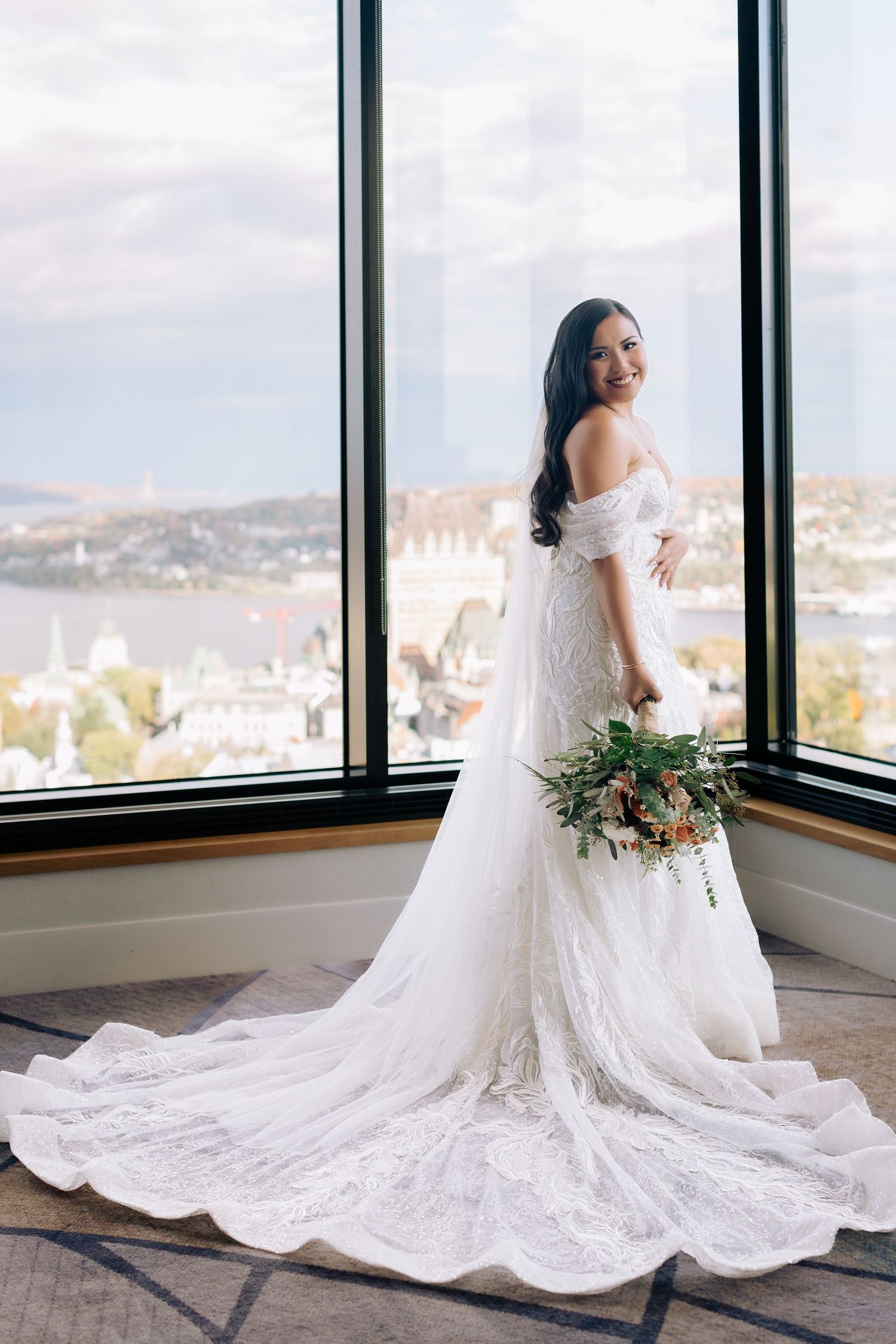 Bride in off-shoulder lace wedding dress poses by window with trailing train and bouquet.