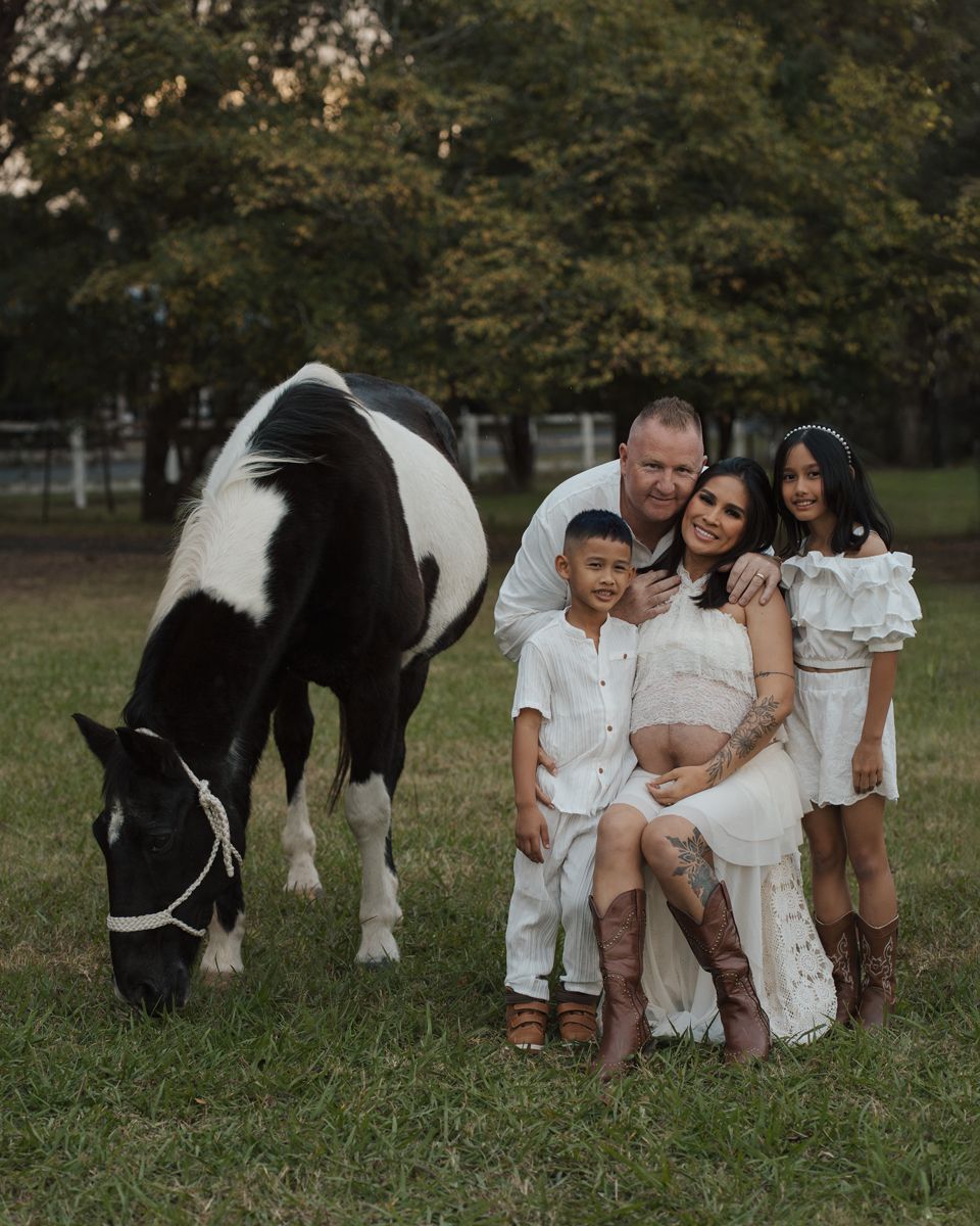 Family maternity photo with parents and two children in white outfits posing beside a black and white horse on a green pasture in Sydney
