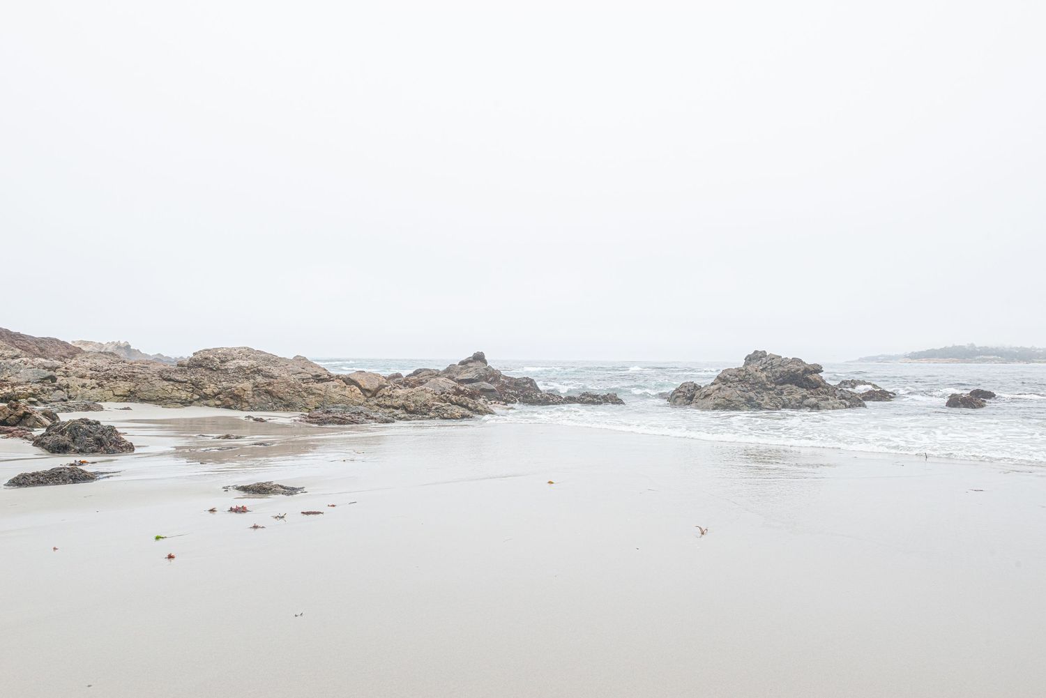 Rocky shoreline on a misty beach with calm waves and gray overcast sky creating a serene coastal landscape.
