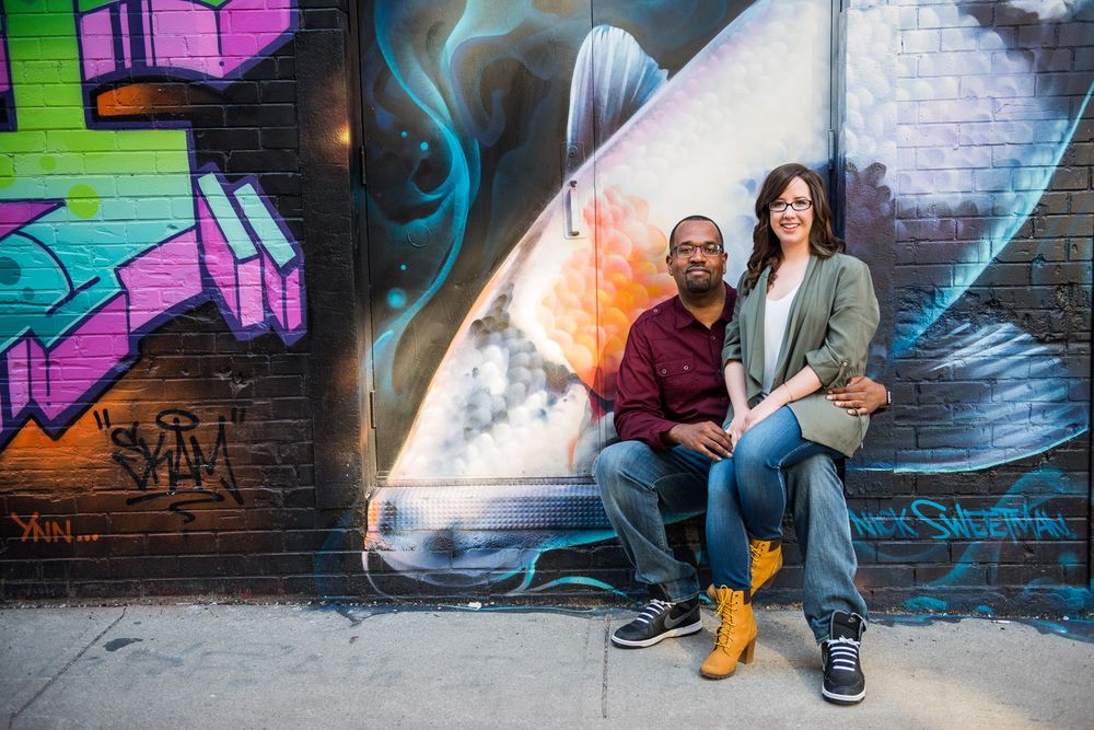 Couple sitting together against a colorful street art mural, woman resting on mans knee in Graffity Alley in Toronto..