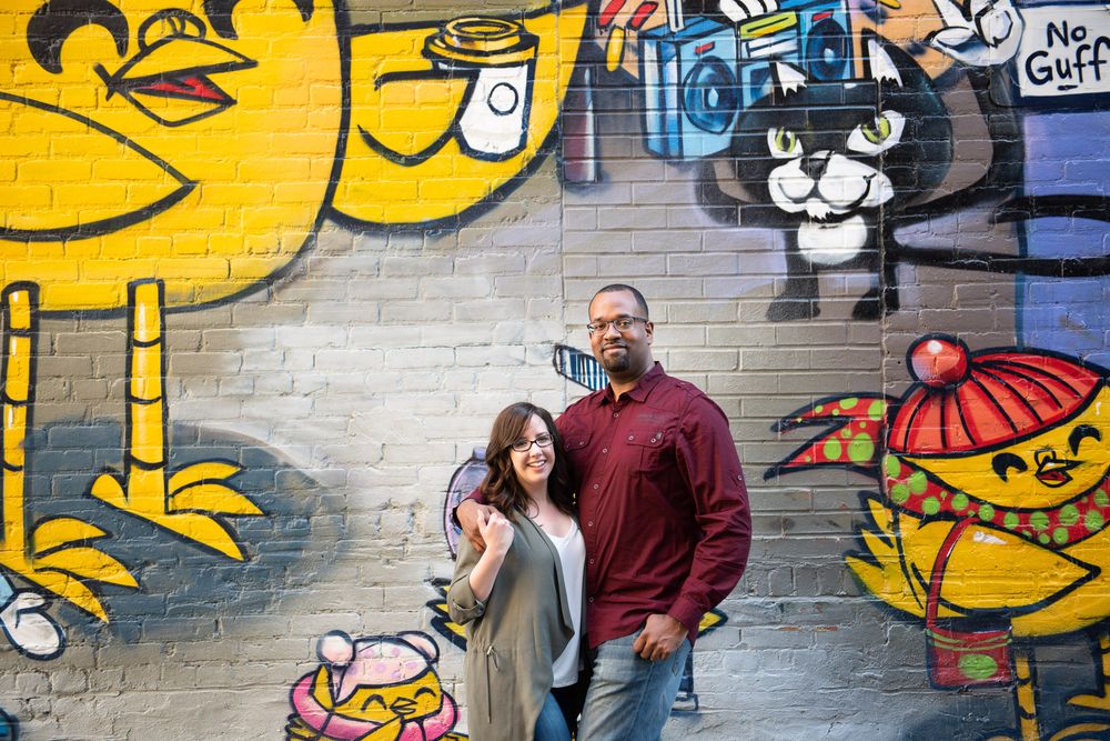 Smiling couple standing before bright cartoon graffiti wall featuring yellow birds and cat characters in Graffity Alley in Toronto..