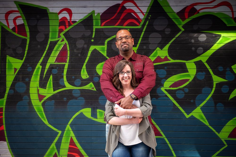 A couple embraces in front of colorful graffiti wall, man hugging woman from behind, both smiling in Graffity Alley in Toronto..