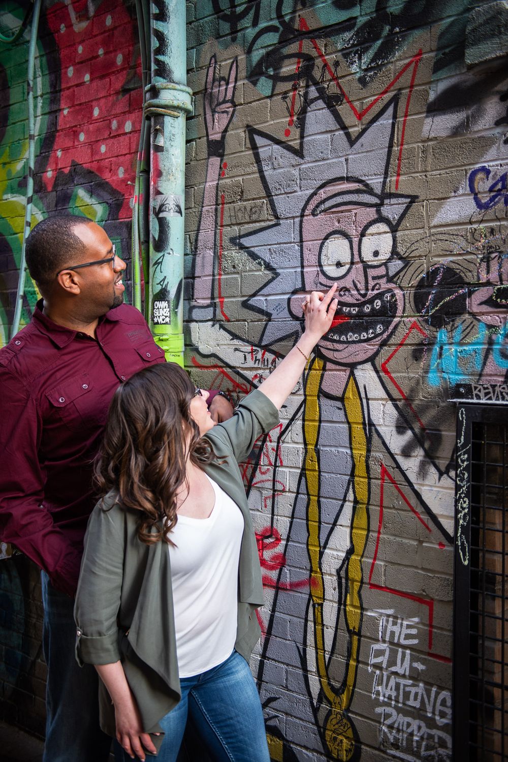 Woman pointing at Rick and Morty graffiti mural on brick wall while man looks on smiling in Graffity Alley in Toronto..