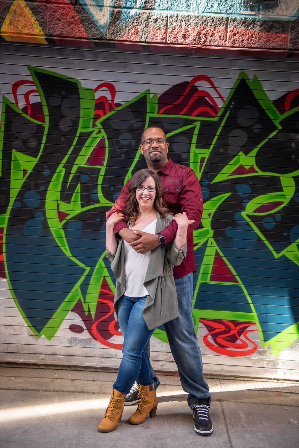 Happy couple posing together in front of vibrant graffiti mural, man embracing woman from behind outdoors in Graffity Alley in Toronto..