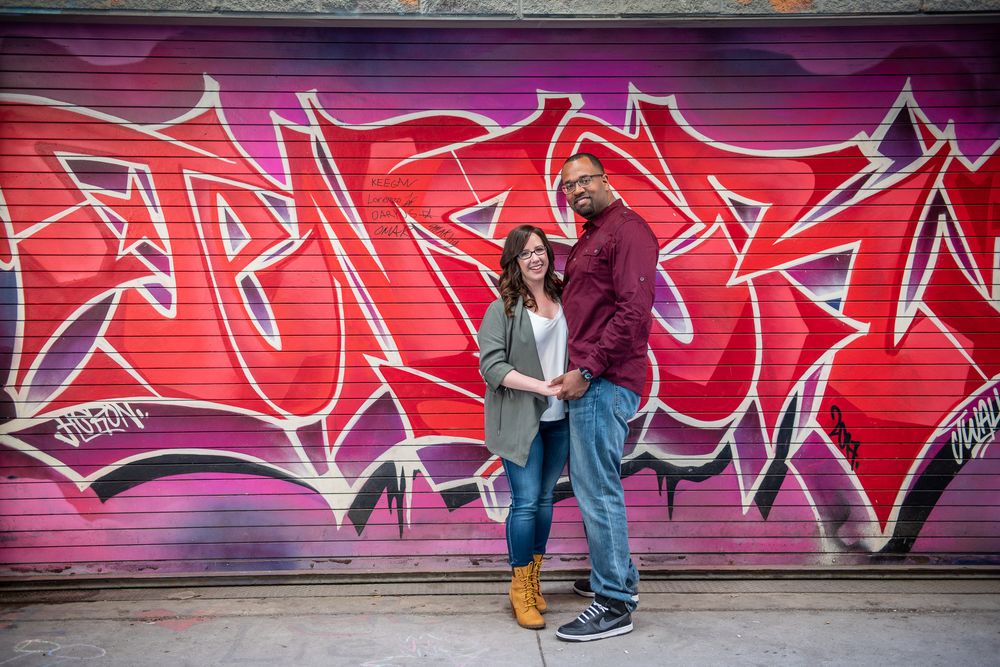 Smiling couple standing together in front of bold pink and purple graffiti wall, holding hands in Graffity Alley in Toronto..
