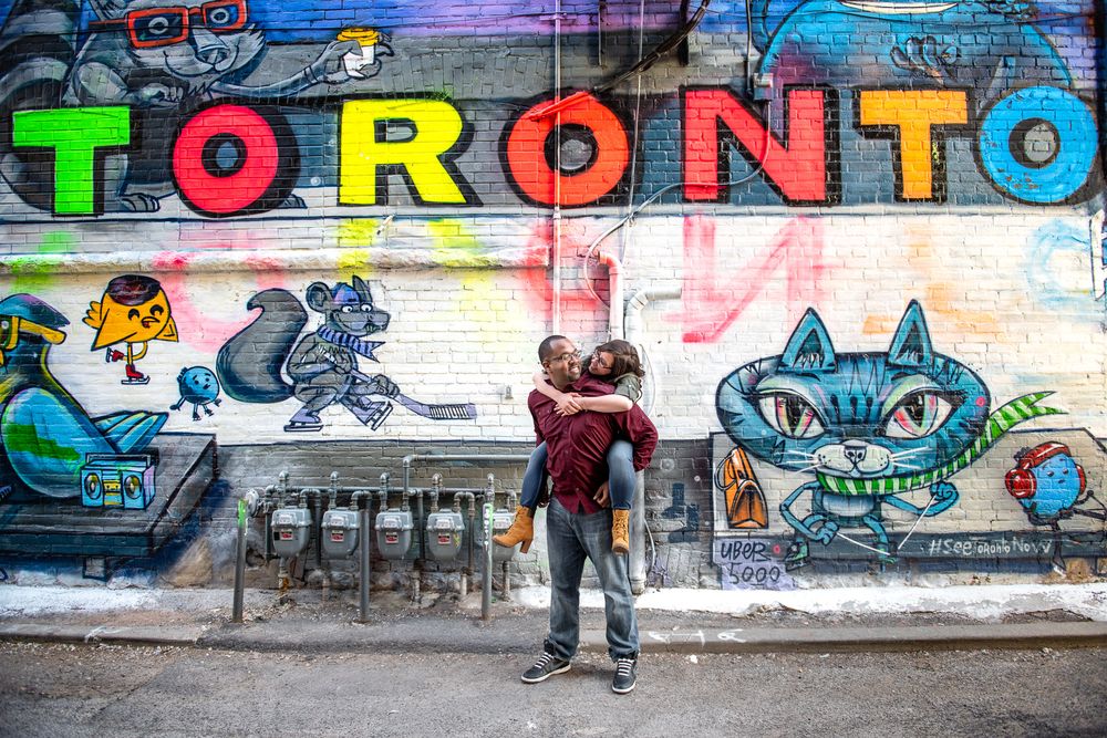 Couple hugging in front of bold Toronto graffiti mural with colorful cartoon characters in Graffity Alley in Toronto..