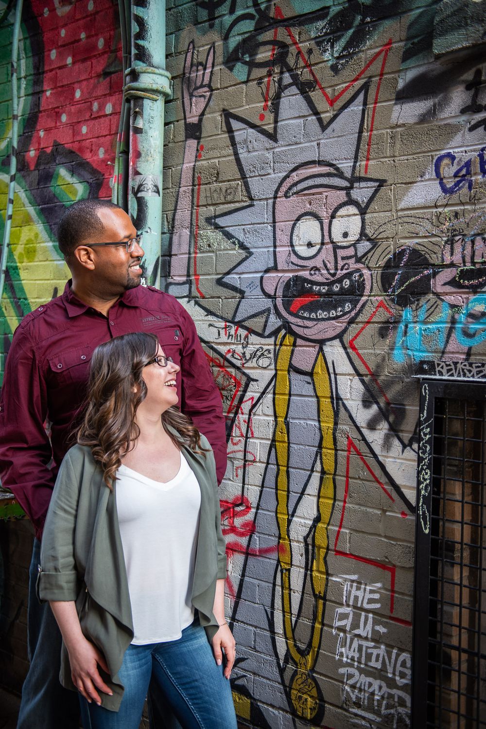 Couple smiling at each other beside Rick and Morty graffiti art on a brick wall in Graffity Alley in Toronto..