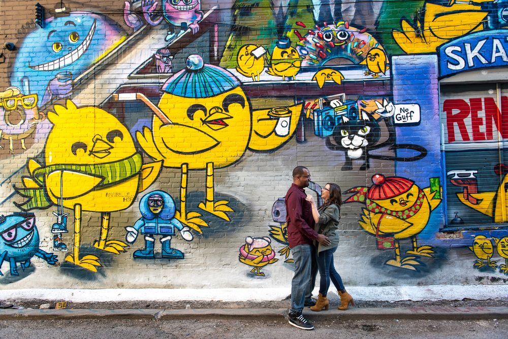 Couple walking past vibrant yellow cartoon character graffiti mural on urban brick wall in Graffity Alley in Toronto..