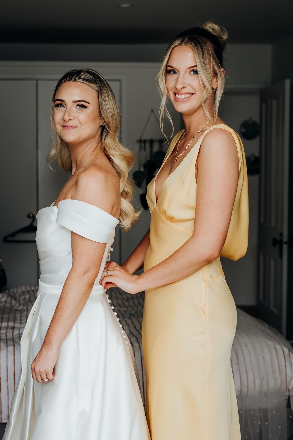 Bridesmaid in yellow satin dress helping bride in off-shoulder white gown get ready for wedding.