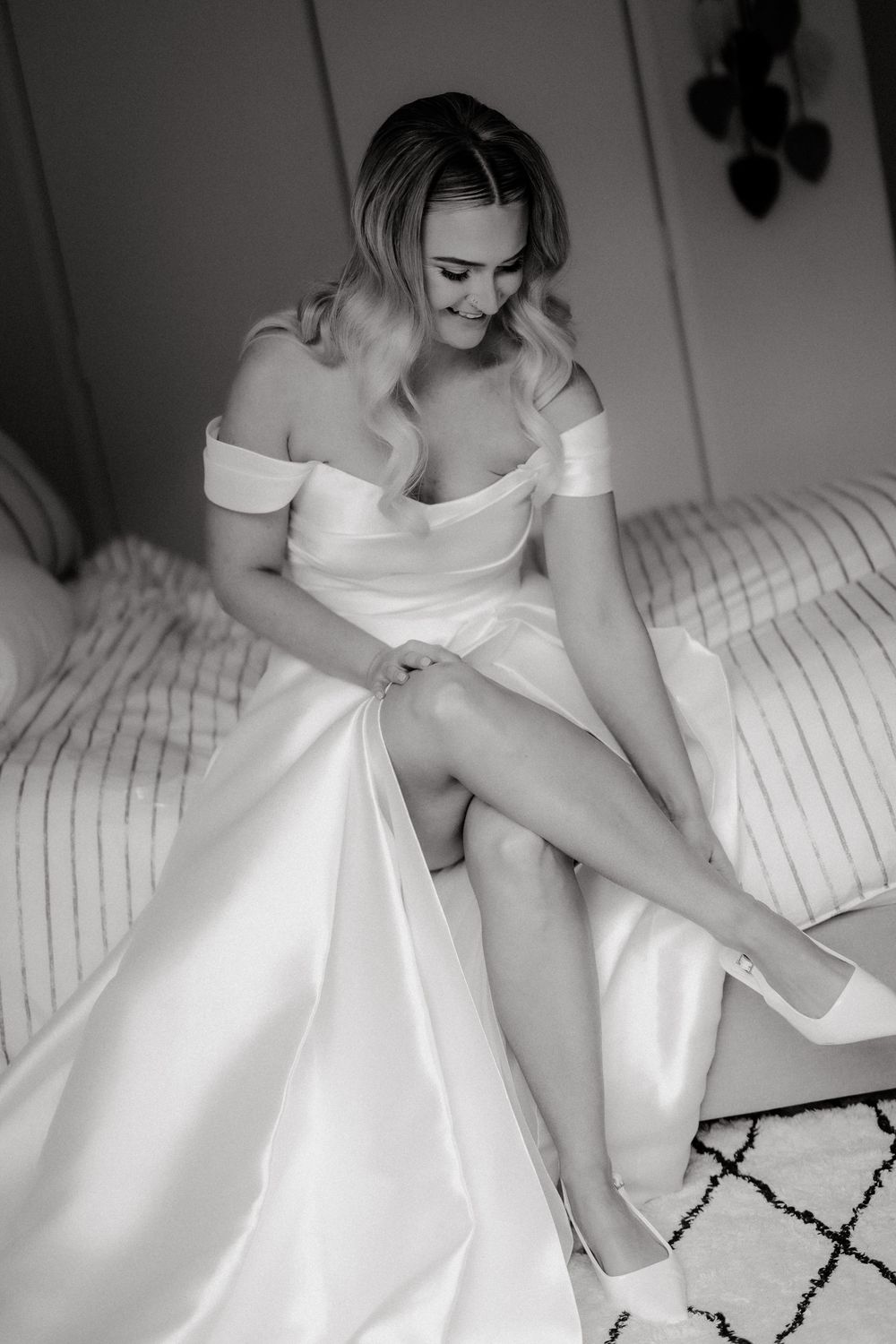Bride in off-shoulder white gown sitting on bed putting on heels, black and white wedding photo.