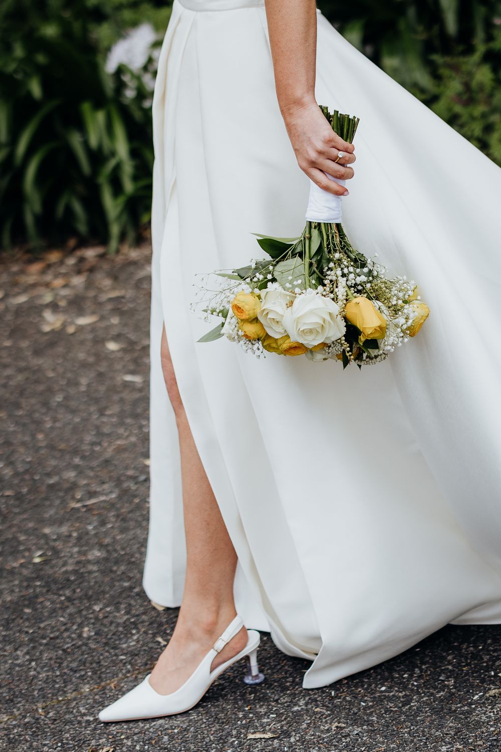 Bride in a white satin gown with a slit holding a yellow and white rose bouquet with white heels.