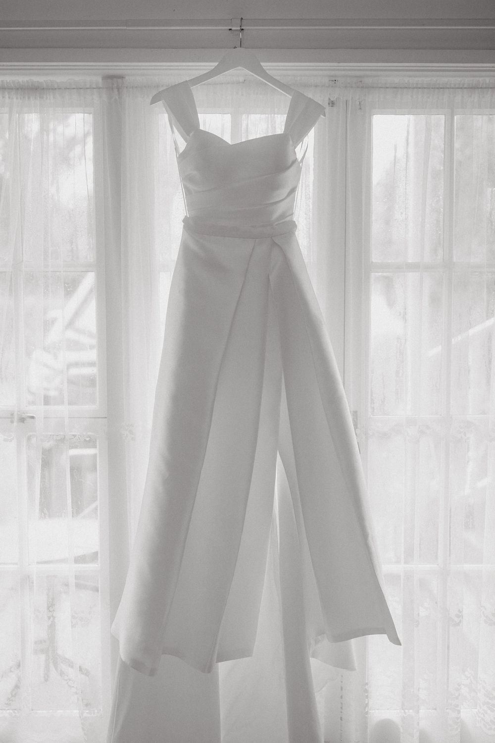 Elegant white bridal jumpsuit hanging on a hanger in front of a bright window with sheer curtains.