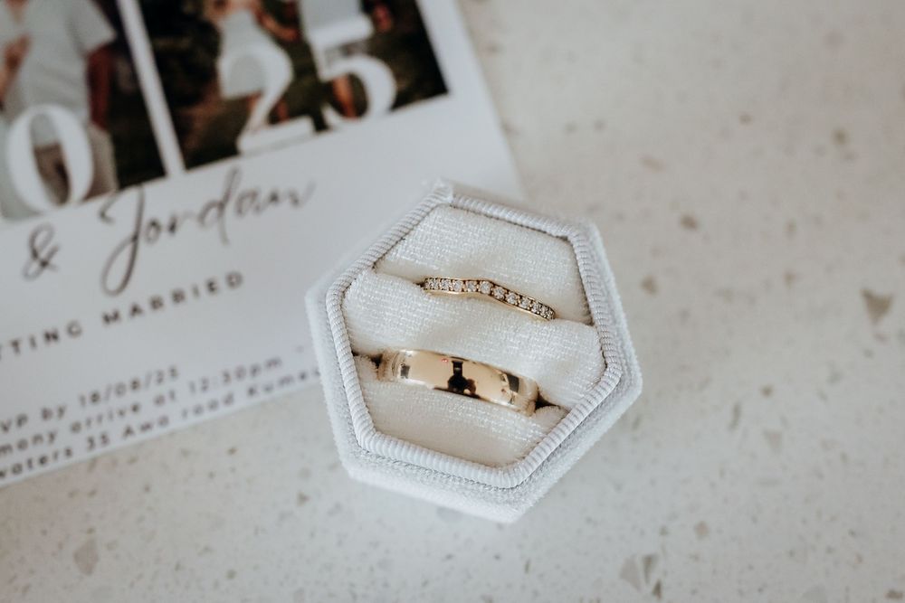 Two gold wedding bands displayed in a white velvet hexagonal ring box beside a wedding invitation card.