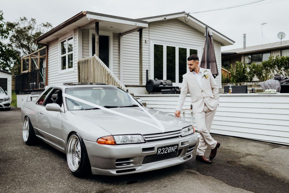 Groom in a beige suit posing beside a silver Nissan Skyline R32 decorated with white wedding ribbons.