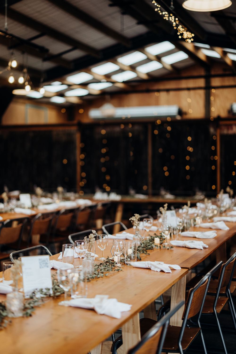 Rustic barn wedding reception with long wooden tables, greenery runners, candles, and fairy lights creating a warm ambiance.