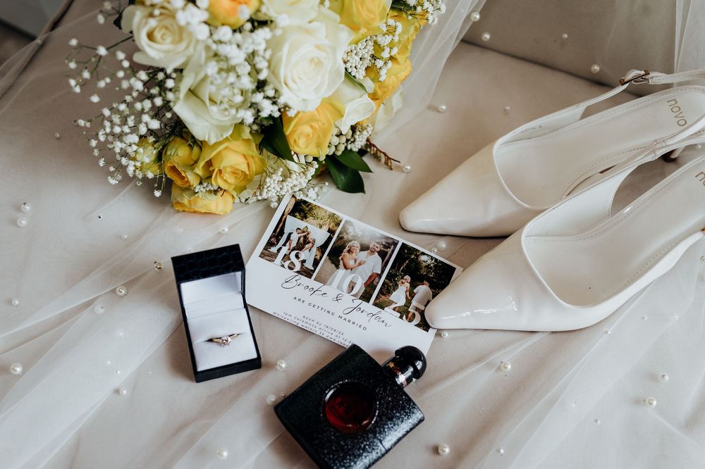 Wedding flat lay featuring yellow roses bouquet, white heels, ring box, perfume, and photo cards.