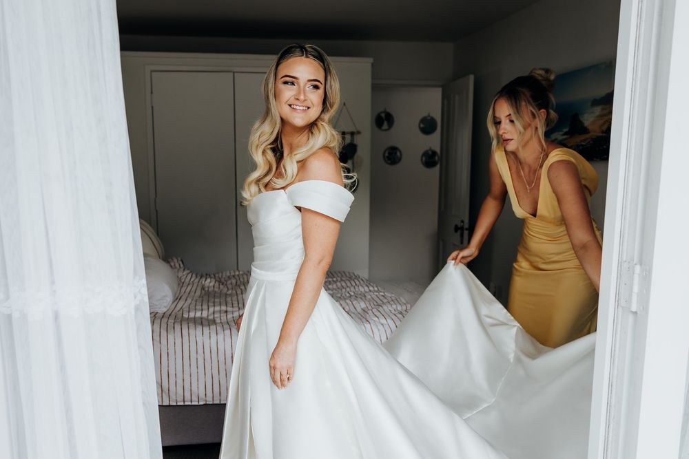 Bride in off-shoulder white wedding gown smiles as bridesmaid in yellow dress adjusts her train.