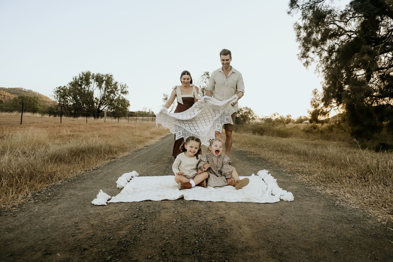 Family of four enjoying a sunny outdoor photo shoot on a dirt road surrounded by golden fields and trees.