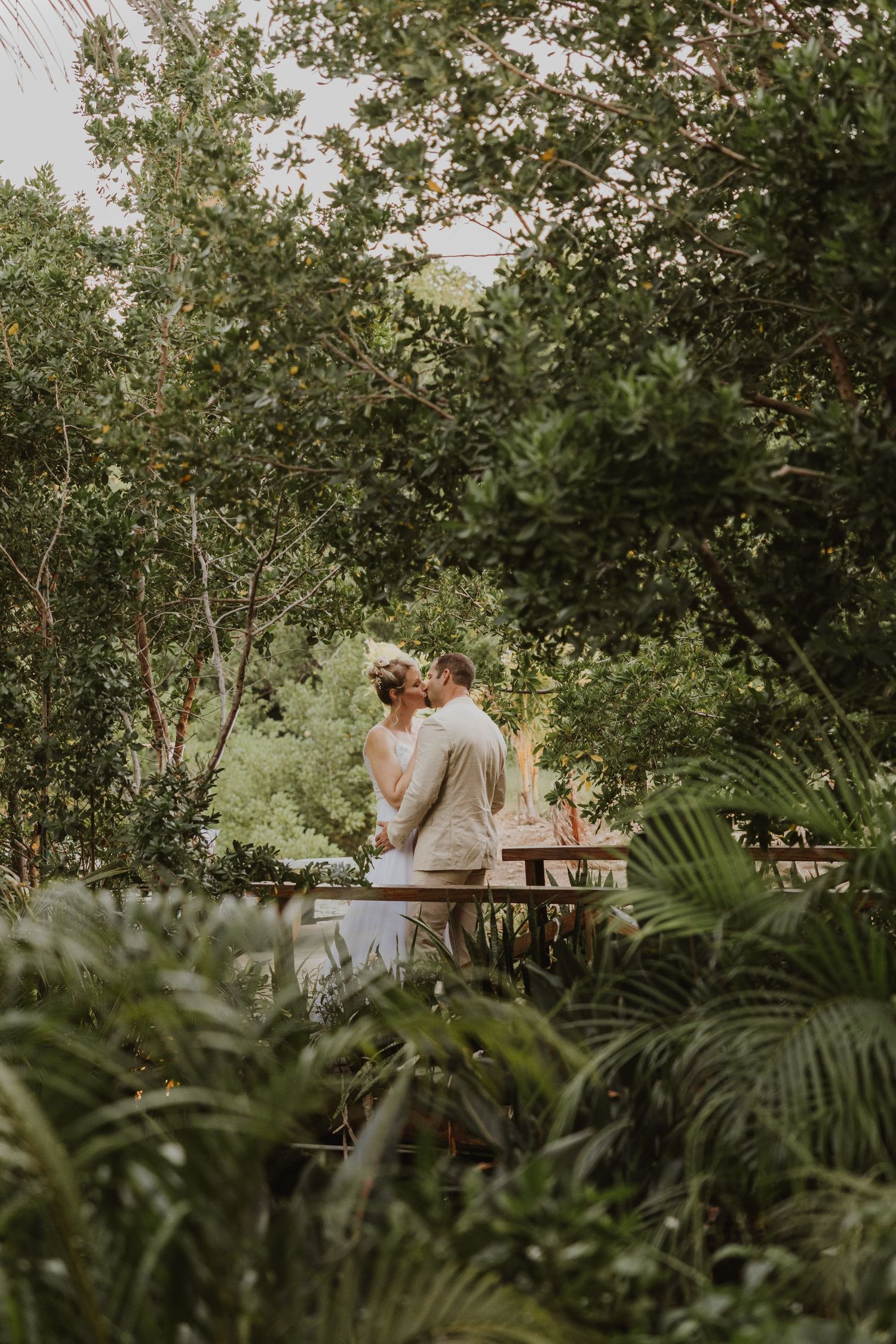 A couple shares a romantic kiss on a wooden bridge surrounded by lush tropical greenery in a garden setting at the aruba ocean villas