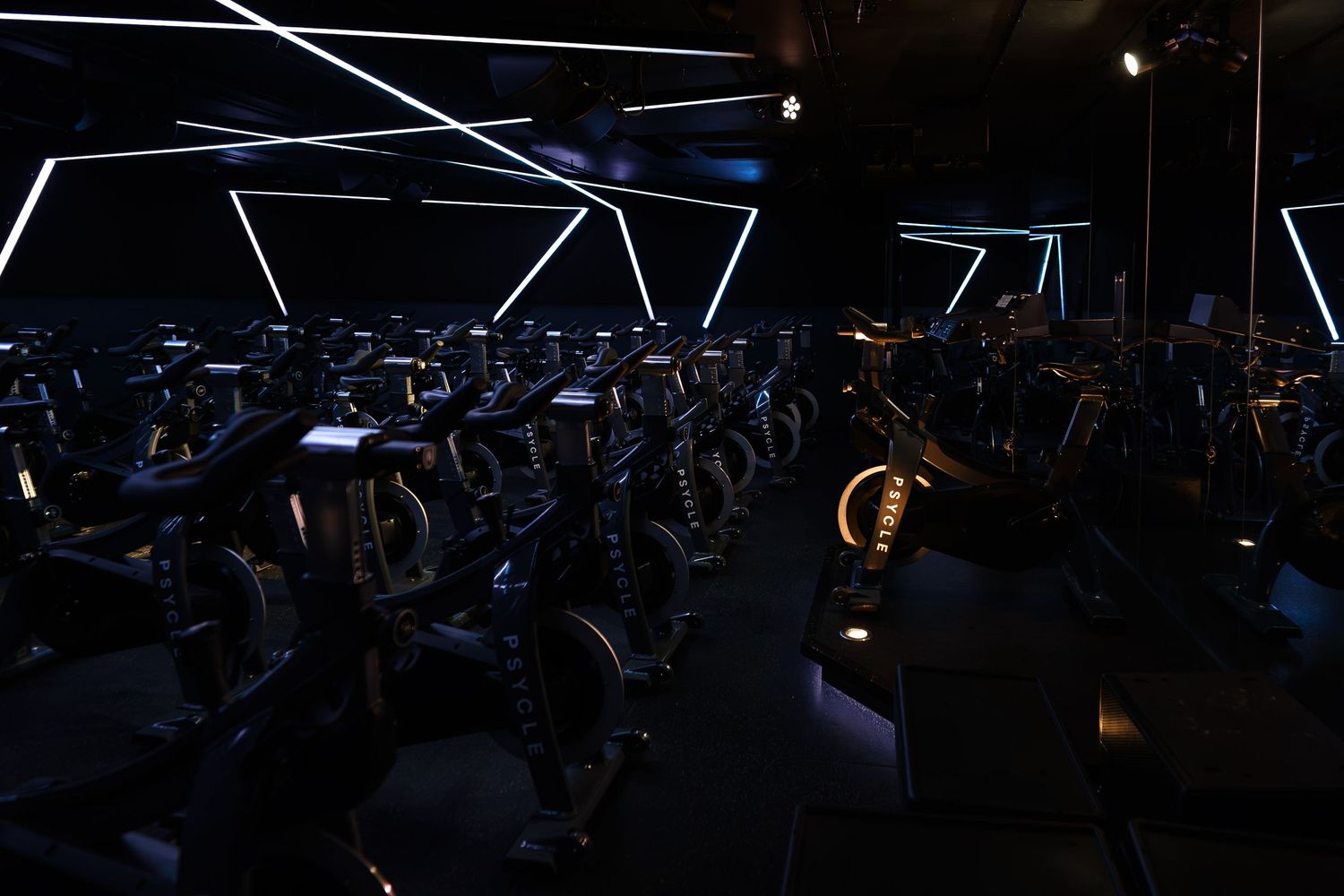 Darkened indoor cycling studio with rows of stationary bikes under geometric neon light patterns on ceiling.