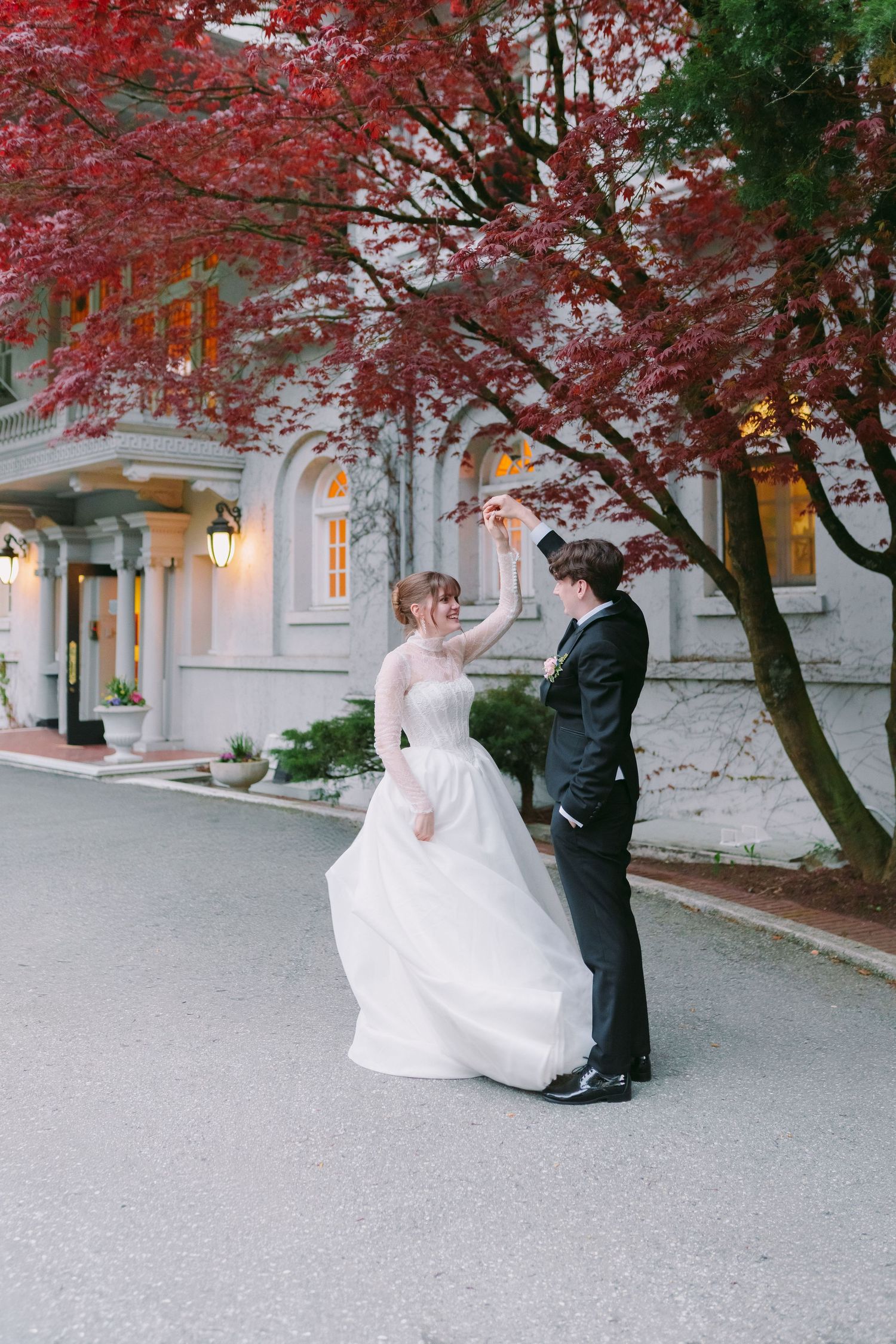Bride in white gown and groom in black tuxedo pose before elegant white mansion with columns and ivy.