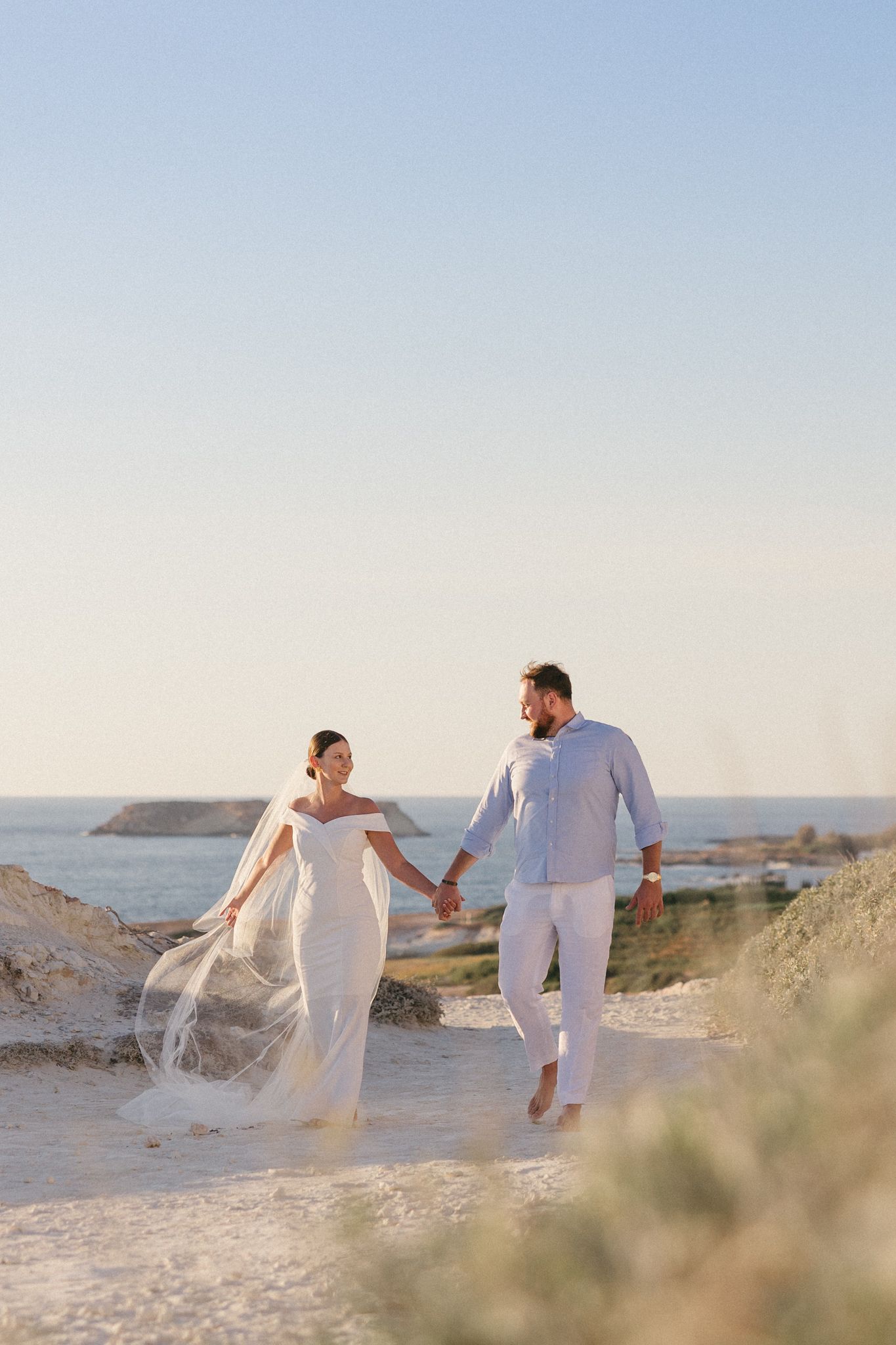 Two people in white outfits walking together on a sandy beach with ocean and cliffs in the background at sunset.