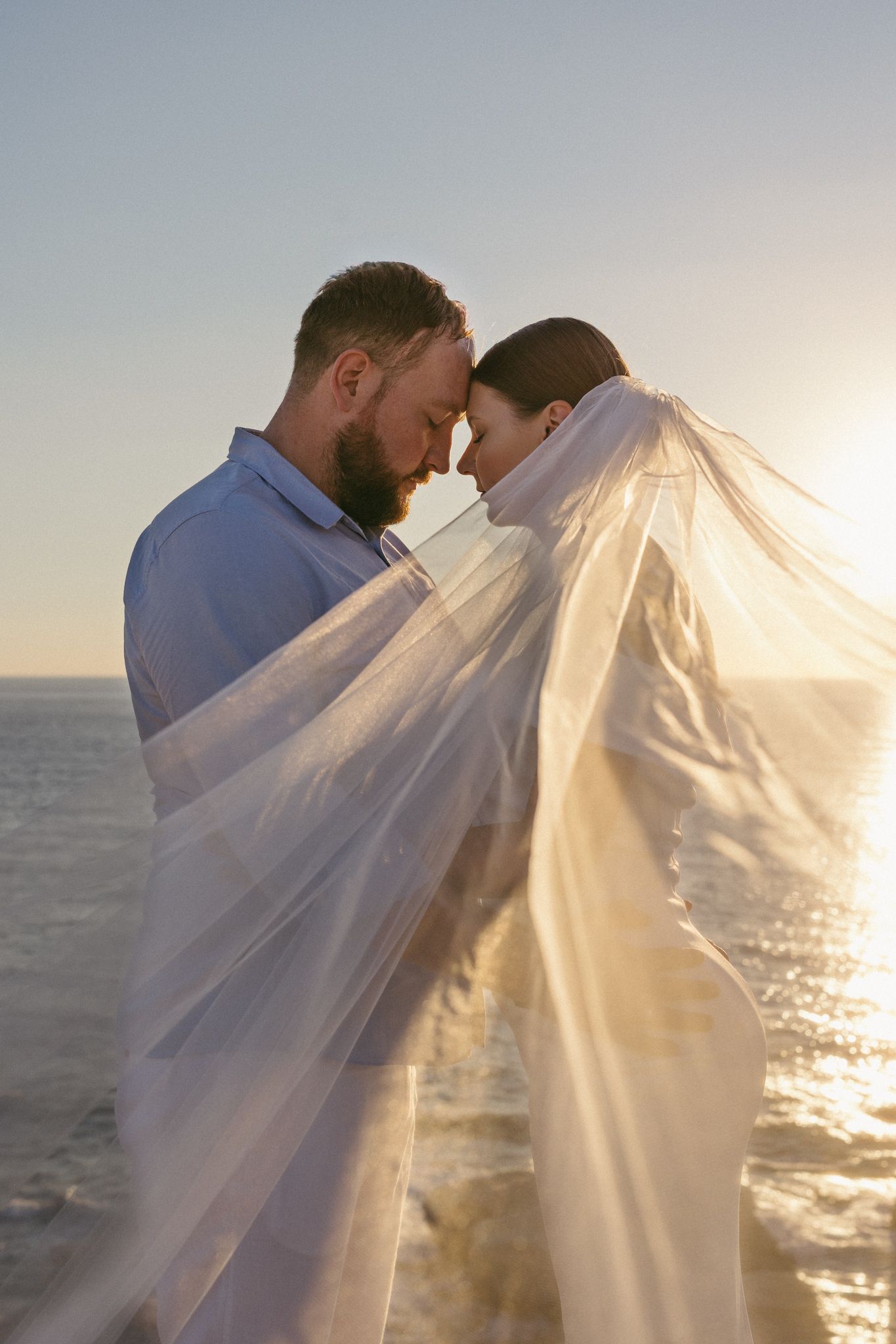 A romantic couple embraces under a flowing veil at sunset by the ocean, creating a dreamy silhouette against the glowing sky.