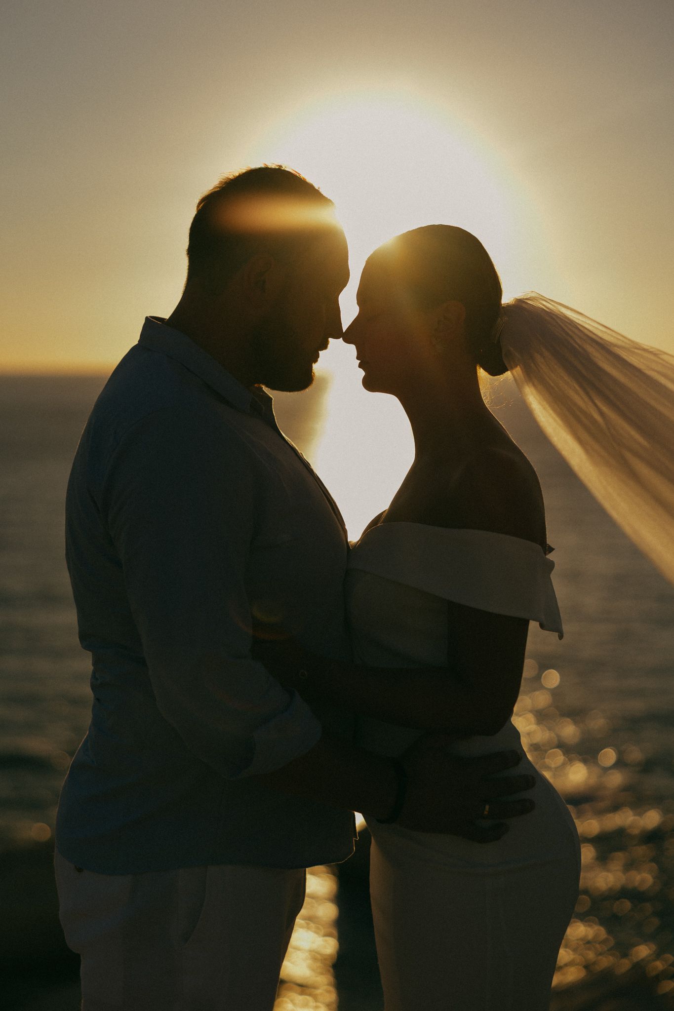 Silhouetted couple embraces at sunset with veil flowing in the breeze against golden ocean backdrop after a wedding in Cyprus.