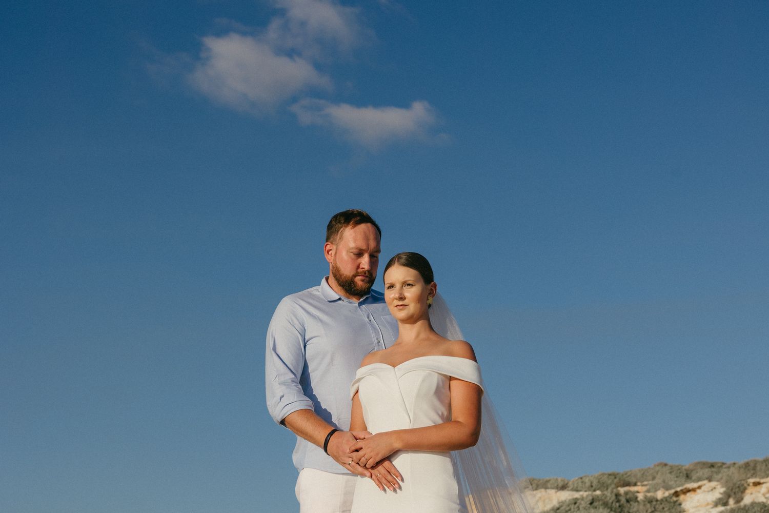 A couple shares tender moments on a beach at sunset against a blue sky and coastal landscape in Cyprus.