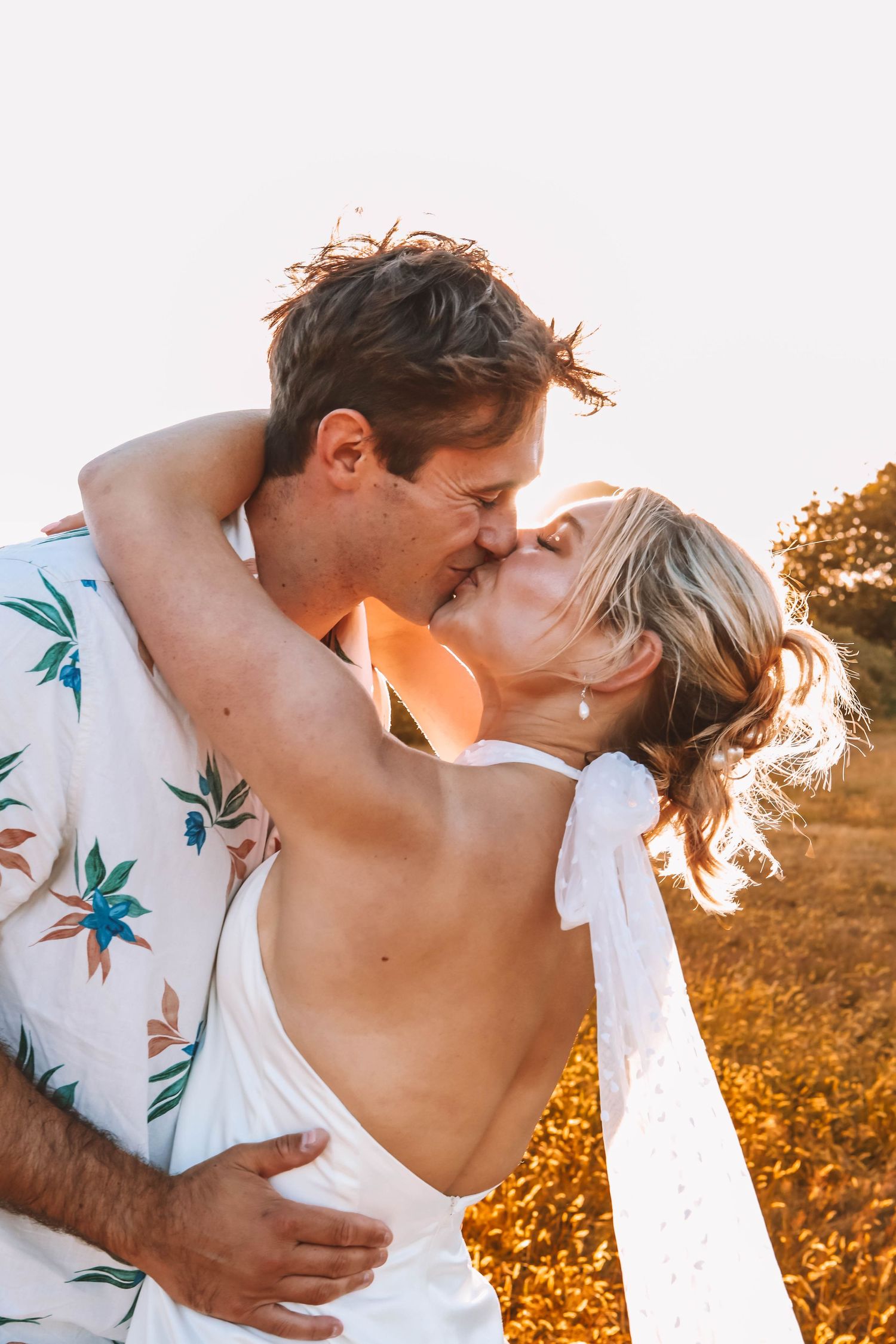 Couple sharing a romantic kiss outdoors at golden hour, woman in white backless dress, man in floral shirt.