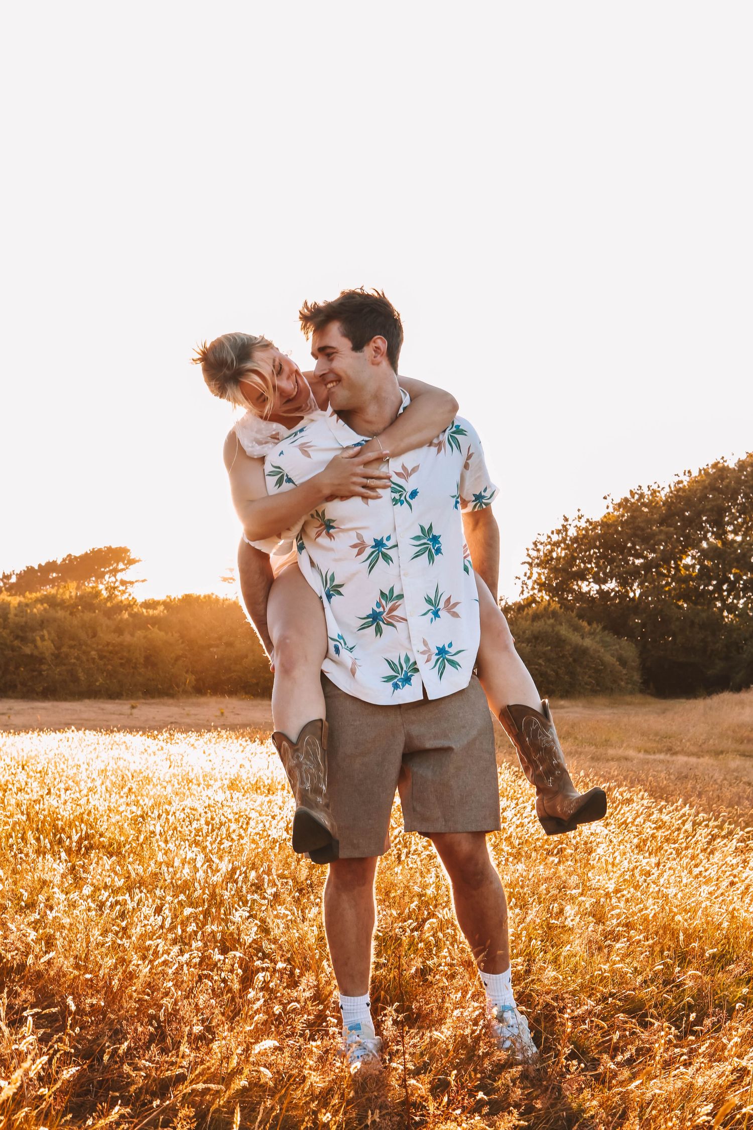A couple shares a joyful piggyback ride in a golden sunlit field, the man wearing a floral shirt and shorts.