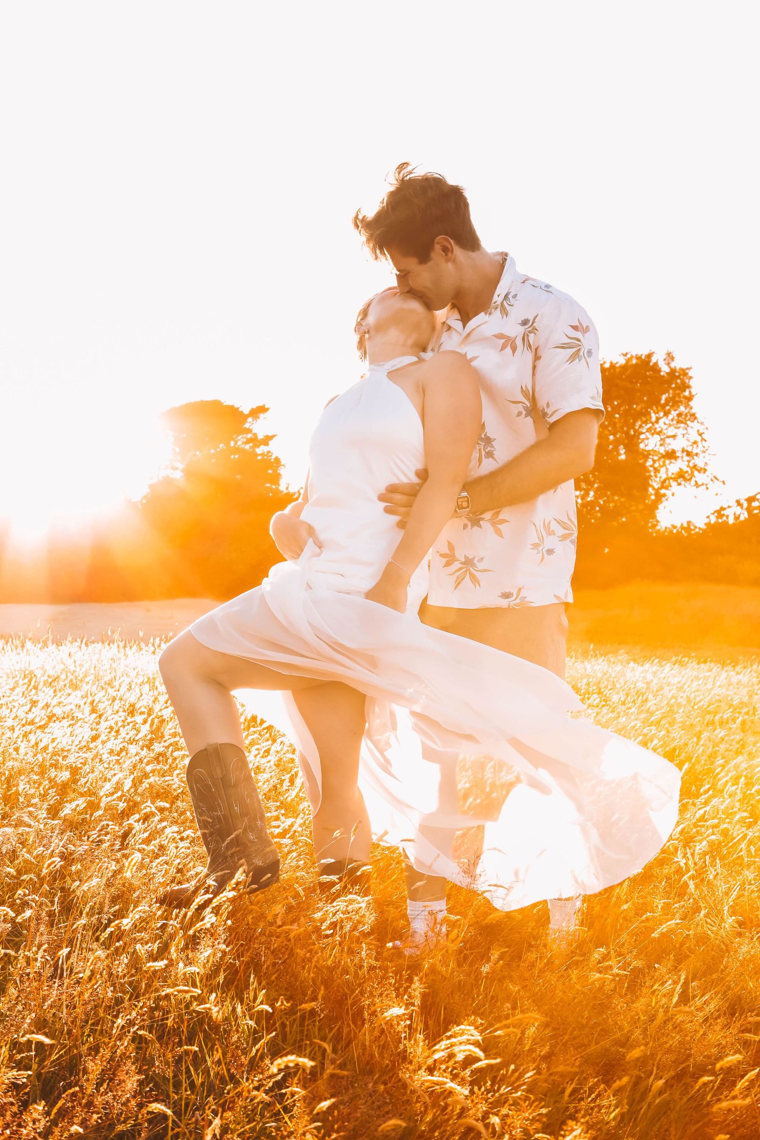 Couple dancing romantically in a golden sunset field, man kissing woman in white dress and boots.