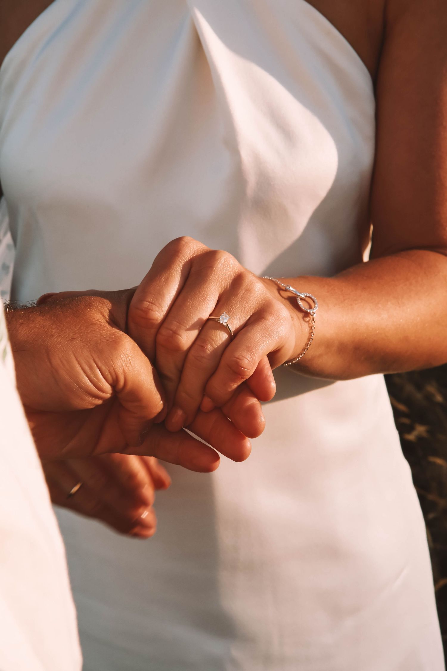 Bride and groom holding hands, showcasing a diamond engagement ring and gold bracelet, wearing a white wedding dress.