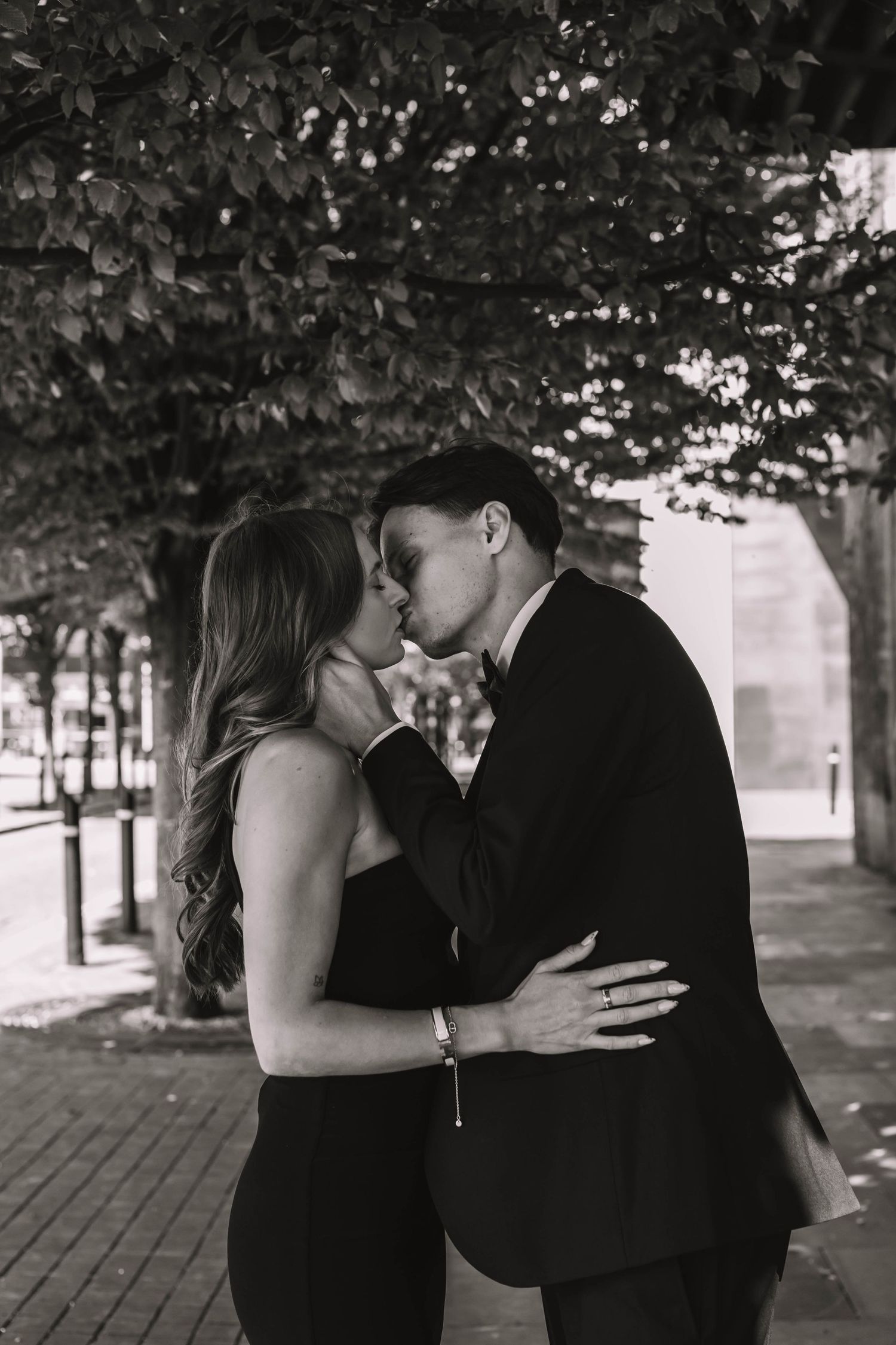 A romantic couple sharing a passionate kiss in a tree-lined walkway, captured in black and white photography.