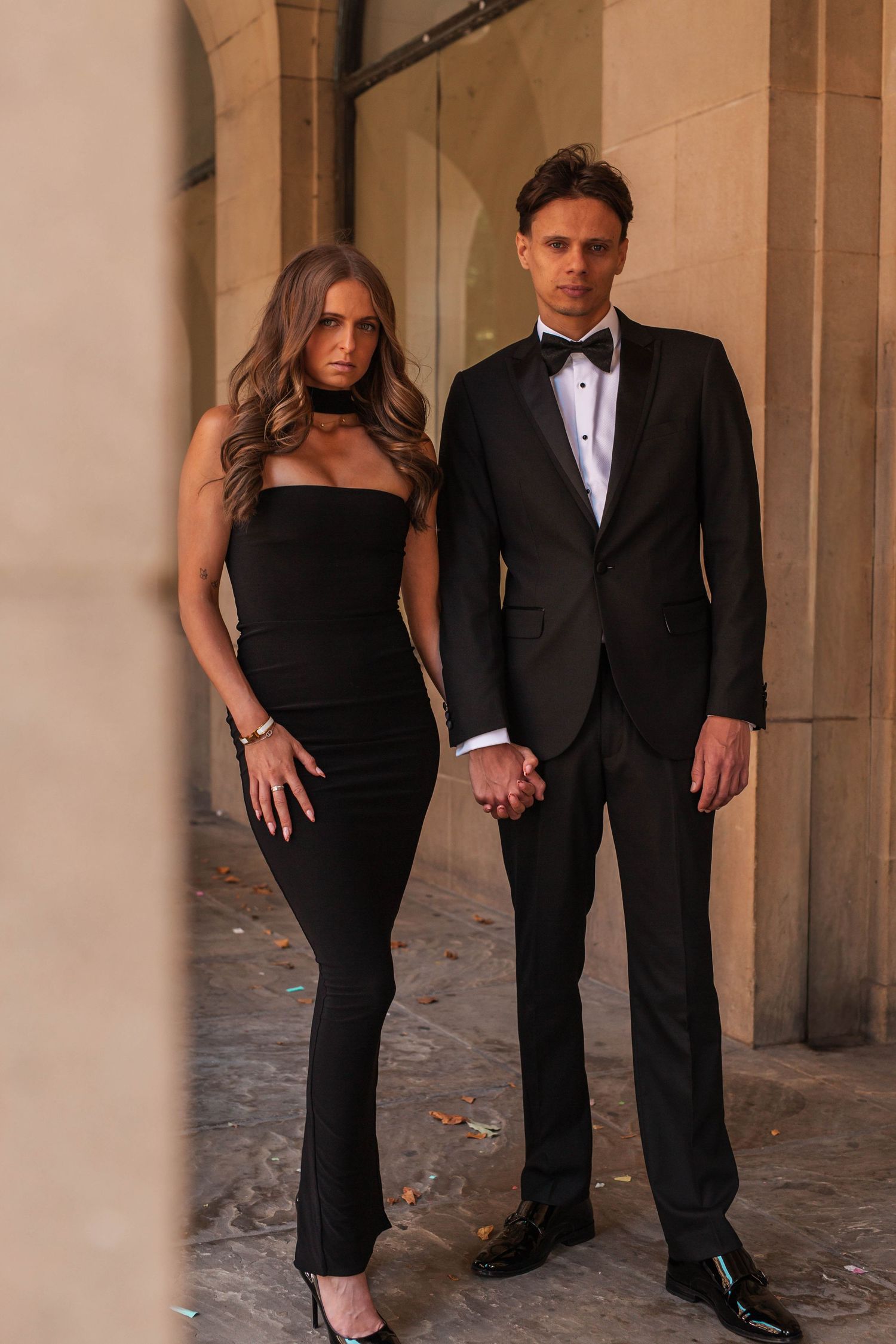 A couple in formal attire poses together against stone pillars in an elegant outdoor setting.