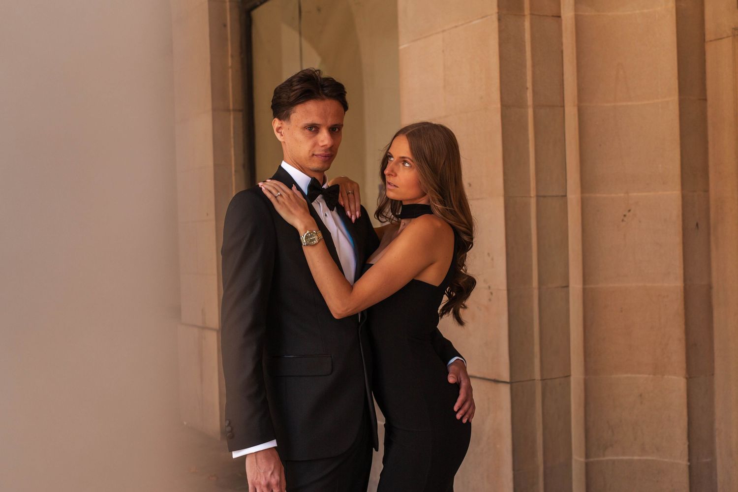 Elegant couple in formal attire, man in black tuxedo and woman in black dress, posing against stone columns.