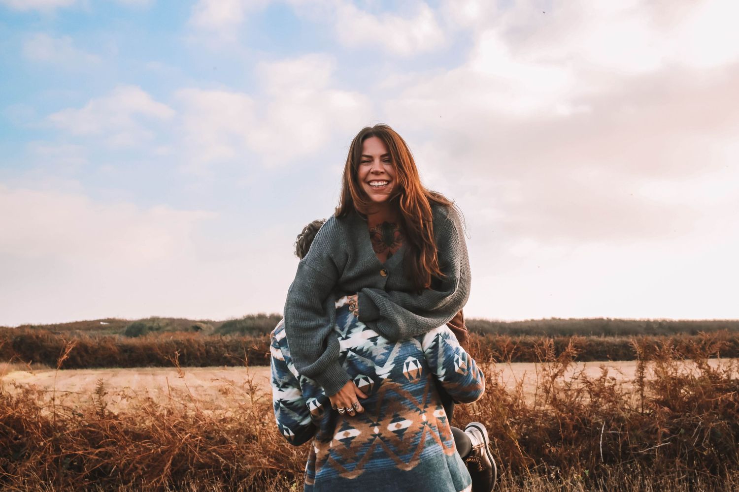 A person with long dark hair wearing a denim jacket poses in a golden field during sunset.