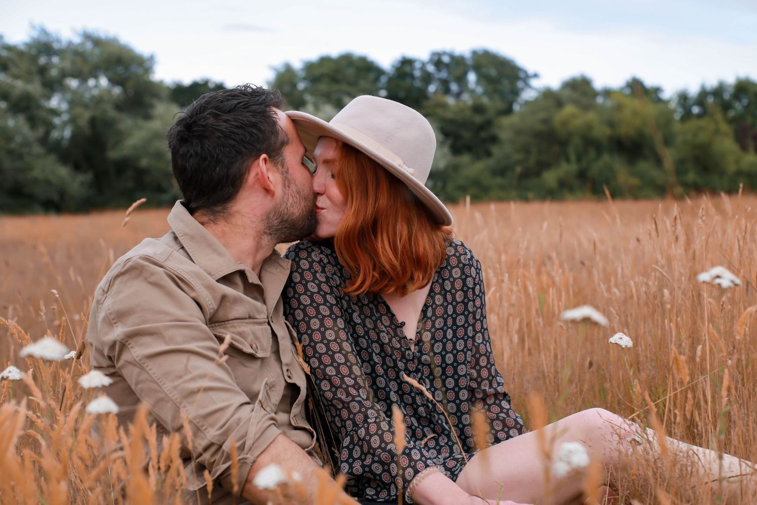 Romantic couple embraces in a golden wheat field during sunset while white dandelion seeds float in the air.