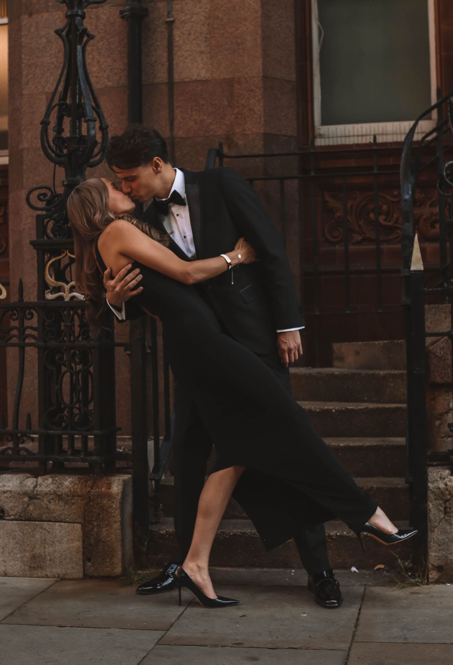 Couple in matching black formal wear sharing a romantic kiss on city steps, woman in elegant gown and heels.