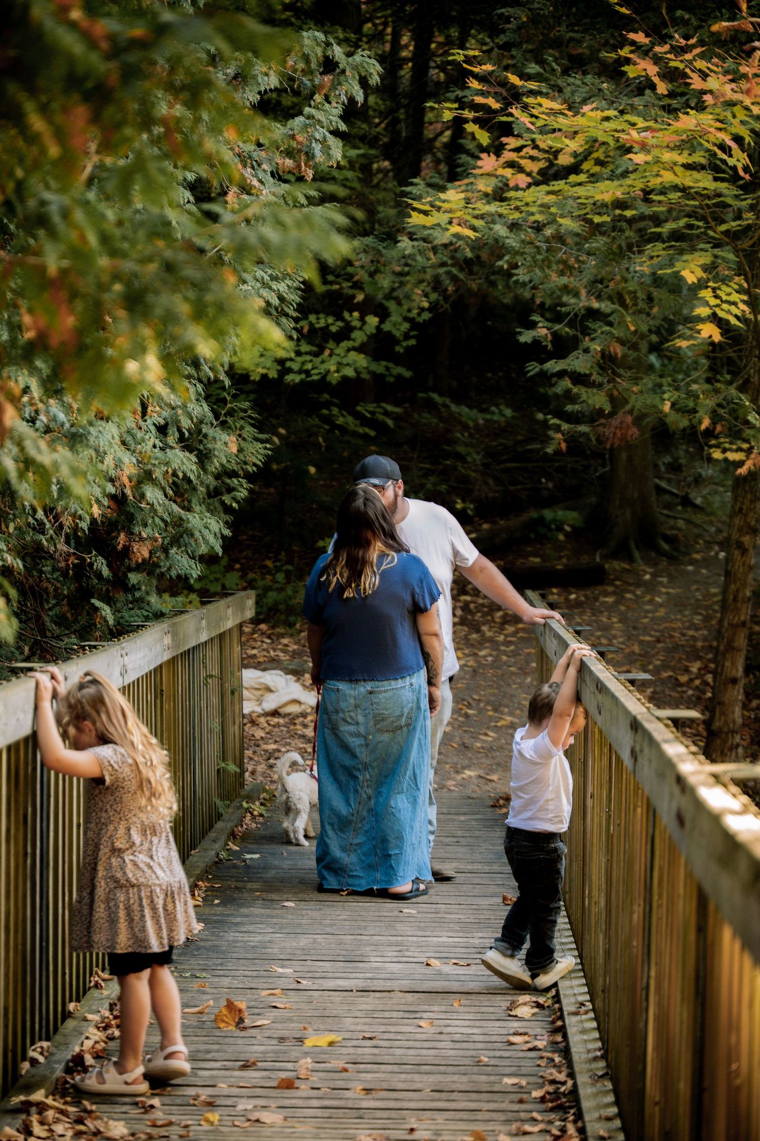 A family walks together on a wooden boardwalk path through autumn trees as fallen leaves scatter around them.