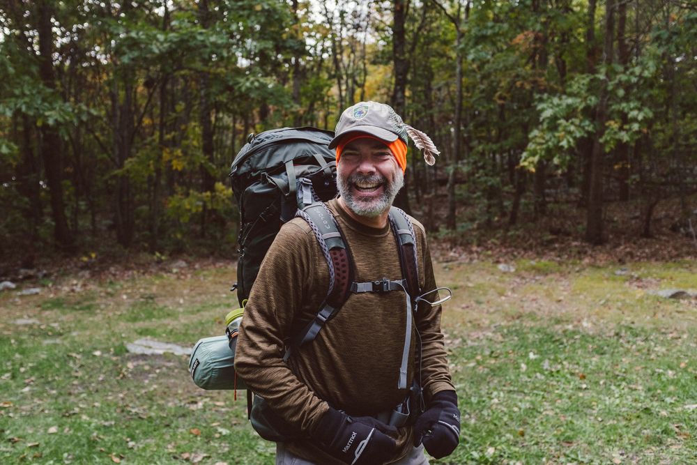 A hunter wearing camouflage and orange gear walks through an autumn forest with a rifle and backpack.