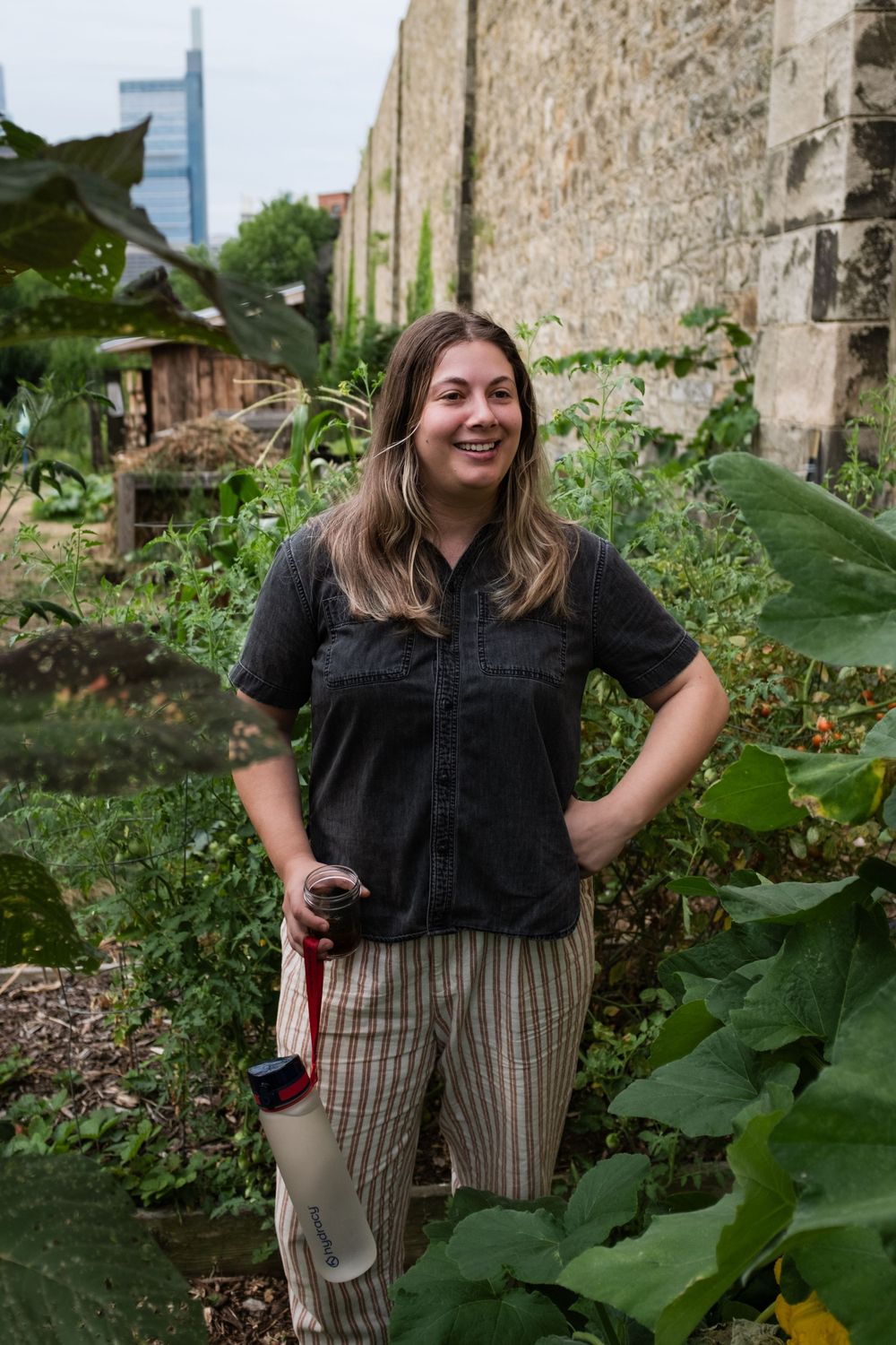 A person in a black shirt and striped pants stands in a lush garden with stone walls and greenery.