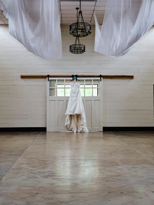 An elegant white wedding dress hangs on a rustic barn door beneath flowing white drapes and iron chandeliers.
