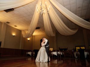 A couple shares their first dance under elegant white ceiling drapes and a crystal chandelier in a ballroom reception space.