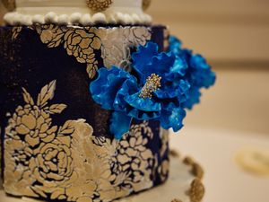 Navy and gold wedding cake decorated with elegant gold patterns and vibrant blue flowers.