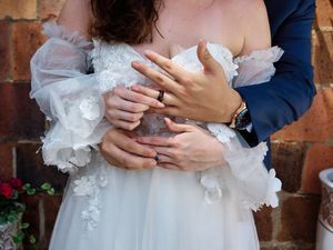 Close-up detail of hands with wedding rings against white dress with delicate floral lace details and brick wall backdrop.