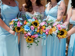Bridesmaids in light blue dresses hold vibrant bouquets with sunflowers and mixed flowers.